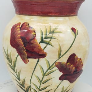Peut inclure: Un vase en céramique avec un corps de couleur crème et un bord rouge foncé. La surface présente des coquelicots rouges peints à la main avec des reflets jaunes, des tiges et des feuilles vertes. Le vase a une forme large et arrondie.