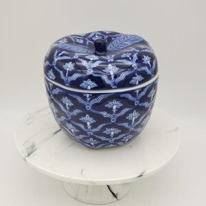 May include: A blue and white ceramic apple-shaped jar with a lid, featuring a floral pattern. The jar sits on a white marble-patterned cake stand. The apple jar has a stem and leaf detail on the lid.
