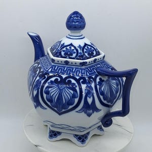 Puede incluir: Una tetera de porcelana blanca con forma hexagonal y complejos patrones florales y geométricos en azul. La tetera incluye una tapa a juego con remate, un pico curvo y un asa azul, todo sobre una base con bordes festoneados.