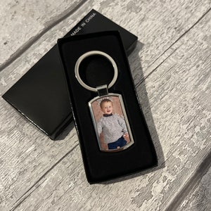 May include: A silver keyring with a rectangular photo insert featuring a young child wearing a grey sweater and blue jeans. The keyring is in a black box with the words "Made in China" printed on the box.