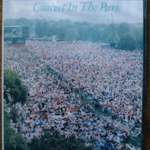 Könnte beinhalten: DVD-Hülle für Paul Simons "Concert In The Park". Das Cover zeigt eine große Menschenmenge in einem Park, mit der Konzertbühne im Hintergrund. Der Text "Paul Simon" steht oben, darunter "Concert In The Park".