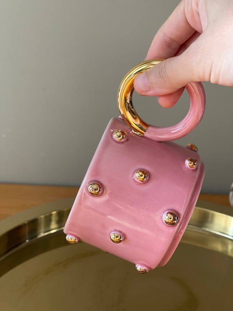 May include: Pink ceramic mug with a gold handle and gold dots. The mug is sitting on a gold tray.