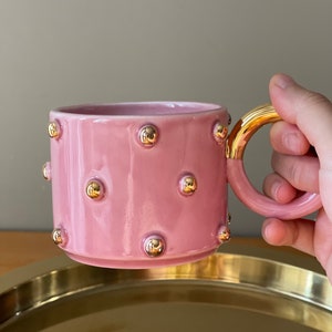 May include: A pink ceramic mug with a gold handle and gold dots around the mug.