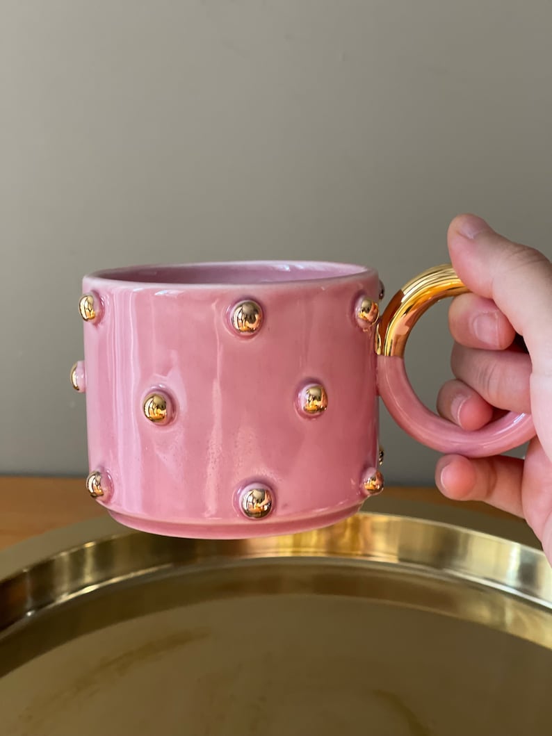 May include: A pink ceramic mug with a gold handle and gold dots around the mug.