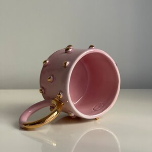 May include: A pink ceramic mug with a gold handle and gold dots around the rim. The mug is empty and sitting on a white surface.