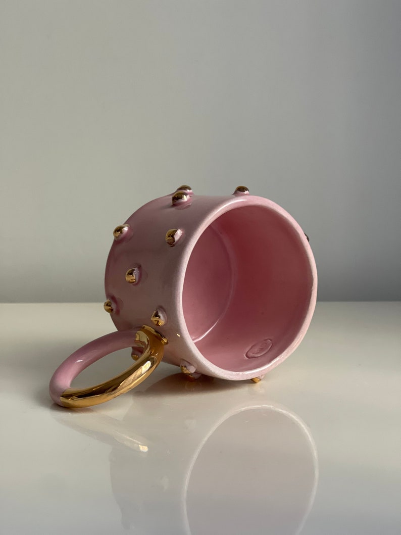 May include: A pink ceramic mug with a gold handle and gold dots around the rim. The mug is empty and sitting on a white surface.