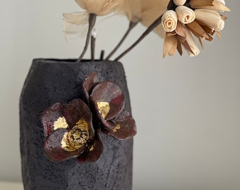 Puede incluir: Un jarrón de cerámica negro con una superficie texturizada y dos flores burdeos con detalles dorados. El jarrón contiene un ramo de flores secas.