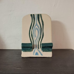 Op de afbeelding: Een witte keramische telefoonstandaard met een abstract ontwerp in blauw en groen. De standaard heeft twee kleine, groene, rechthoekige uitsteeksels aan de onderkant om een telefoon te houden.