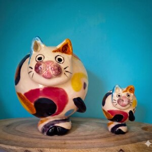 Peut inclure: Deux figurines de chat en céramique. Le grand chat est blanc avec des taches colorées rouges, jaunes et bleues. Le petit chat a des couleurs similaires. Les deux ont des pattes et des moustaches noires, avec des nez roses et des fonds bleus.
