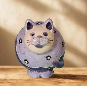 May include: A ceramic cat figurine in shades of lavender and white. The cat has a round body with black flower accents and a curled tail. The cat's face is white with purple accents on the ears and nose.