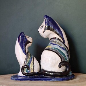 May include: A ceramic cat sculpture set featuring two stylized cats. The cats are white with blue, black, and multicolored accents. The cats sit on a blue base, with curled black tails. The sculpture has a whimsical, artistic design.