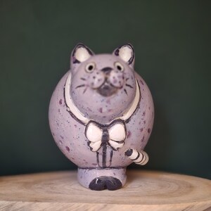 Puede incluir: Una figura de gato de cerámica redonda con un esmalte moteado de color morado claro. El gato presenta un cuello blanco, una pajarita blanca y detalles pintados en morado oscuro en la cara, las orejas y las patas. Una pequeña cola rayada se curva.