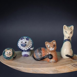 May include: Four ceramic cat figurines in various poses and colors. One small round cat, one round cat with a blue patterned collar, one reclining brown cat with black stripes, and one tall cream-colored cat with blue accents on its neck and tail.