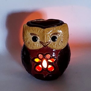 Può includere: Un portacandele in ceramica a forma di gufo con corpo marrone scuro e viso color cuoio. Il gufo presenta grandi occhi neri e un intaglio a forma di fiore sul petto, che emana una calda luce arancione proveniente da una fiamma interna.
