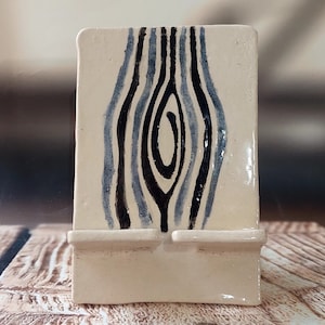 Puede incluir: Un soporte de teléfono de cerámica blanca con un diseño abstracto en negro y azul. El soporte tiene dos ranuras para sujetar teléfonos.