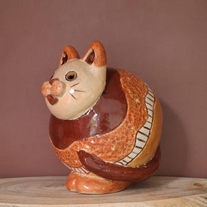 Puede incluir: Figura de gato de cerámica en tonos beige, marrón y naranja. El gato tiene un cuerpo redondeado con un patrón texturizado y una cola enroscada. La cara del gato tiene detalles, incluyendo bigotes y orejas.
