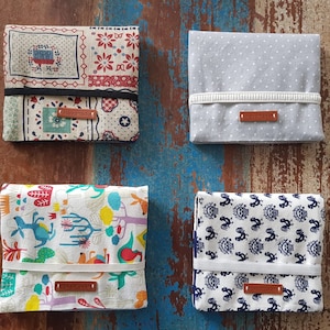 Könnte beinhalten: Vier verschiedene Gewebetuchhalter aus Stoff mit einem braunen Lederlabel. Die Halter haben verschiedene Muster, darunter ein Blumenmuster, einen weißen Hintergrund mit bunten Tieren und Bäumen, einen grauen Hintergrund mit weißen Punkten und einen weißen Hintergrund mit blauen und weißen Hahnmotiven.