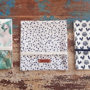 Könnte beinhalten: Drei Stoff-Lebensmittelverpackungen mit verschiedenen Mustern. Die erste Verpackung hat ein Aquarell-Design in Grün-, Blau- und Rosatönen. Die zweite Verpackung hat einen weißen Hintergrund mit kleinen blauen Blumenmustern. Die dritte Verpackung hat einen weißen Hintergrund mit einem sich wiederholenden Muster aus blauen Hähnen.