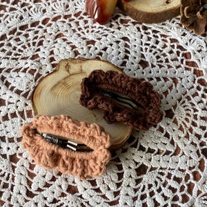 Op de afbeelding: Twee handgemaakte gehaakte haarclips. Eén is lichtbruin, de andere is donkerder bruin. De clips liggen op een houten schijf, op een wit kanten kleedje. Decoratieve stenen en een dennenappel staan op de achtergrond.