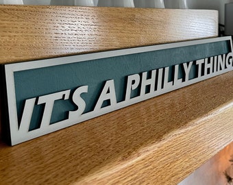 May include: A teal and white wooden sign with the text "IT'S A PHILLY THING" in white letters. The sign is mounted on a wooden shelf.