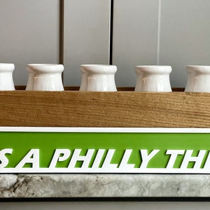May include: A wooden box with five white ceramic milk bottles and a green sign that reads "IT'S A PHILLY THING" in white letters.