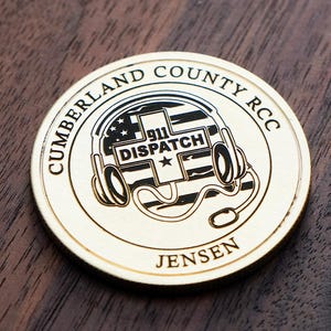 May include: A gold-coloured coin with the text "CUMBERLAND COUNTY RCC" around the edge. The centre features a design of a headset with the words "911 DISPATCH" and a star. The name "JENSEN" is at the bottom. The coin is on a dark wood surface.