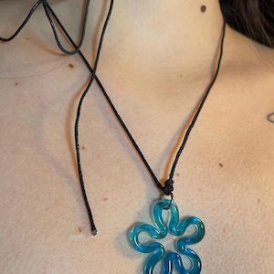 Peut inclure: Un collier avec un pendentif en forme de fleur bleue et un cordon noir. Le pendentif floral est bleu translucide avec un motif tourbillonnant. Le cordon du collier s'attache dans le dos.
