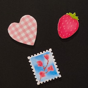Peut inclure: Trois autocollants décoratifs sur fond noir. Un cœur rose et blanc à carreaux, une fraise rouge avec le dessus vert et un timbre-poste avec un motif floral et un fond de ciel bleu.