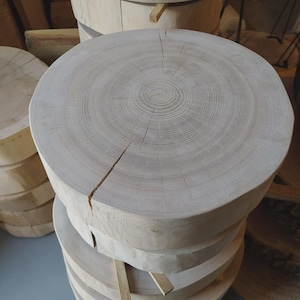Può includere: Una pila di lastre di legno rotonde di colore chiaro, probabilmente sezioni trasversali di alberi, con anelli di crescita e crepe naturali visibili. Il legno ha una finitura chiara e naturale, che suggerisce un'estetica rustica o incompiuta.