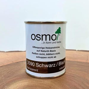 Könnte beinhalten: Eine zylindrische Dose Holzbeize von Osmo, beschriftet mit "3590 Schwarz / Black transparent". Die Dose ist weiß mit goldenem Rand und trägt das Osmo-Logo und Text auf Deutsch. Die Dose steht auf einer Holzoberfläche.