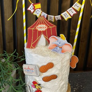 May include: A white cake decorated with a red and gold circus tent, a Dumbo the Elephant character, a peanut-shaped cookie, and a ticket with the text "Circus Amazing Show". The cake is topped with a yellow and white striped banner that reads "Scarlett".