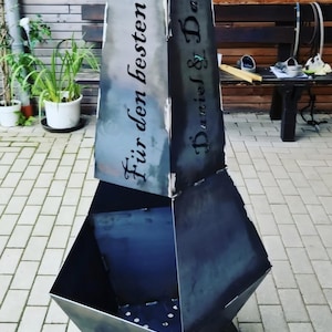 Puede incluir: Un brasero de metal oscuro con un diseño geométrico. El brasero tiene una base rectangular y una sección superior alta e inclinada. El texto en la sección superior dice "Für den besten Papa" y "Daniel & Damien".