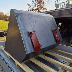 Könnte beinhalten: Eine große, graue Metallschippe mit trapezförmiger Form, die auf Holzbalken ruht. Zwei rote Metallhalterungen sind vorne angebracht. Die Schaufel ist wahrscheinlich für die Landwirtschaft oder den Bau bestimmt.
