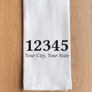 May include: A white rectangular kitchen towel featuring the black printed text 12345 followed by the phrase Your City, Your State in a serif font. The fabric has a visible woven texture and is folded into a neat, vertical rectangular shape.