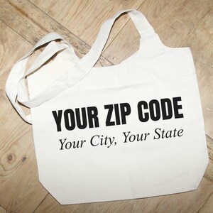 May include: A white canvas tote bag with two shoulder straps featuring the black printed text "YOUR ZIP CODE" in a bold sans-serif font, followed by the text "Your City, Your State" in a smaller, italicized serif font centered on the front.