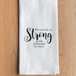 May include: White kitchen towel with the text "We are only as strong as the girlfriends we have" in black script. The towel is folded and placed on a light brown wooden surface.