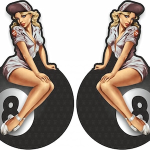 Op de afbeelding: Twee pin-up stijl illustraties van vrouwen in witte outfits en petten, zittend op zwarte en witte 8-ball biljartballen.