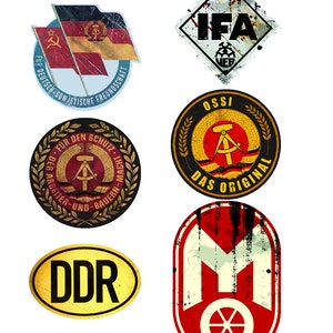 Op de afbeelding: Zes vintage-stijl, verweerde, ronde en ruitvormige stickers met verschillende tekst en symbolen. De stickers bevatten de woorden "DDR", "IFA", "OSSI", "DAS ORIGINAL", "FÜR DEUTSCH-SOWJETISCHE FREUNDSCHAFT" en "FÜR DEN SCHUTZ DER ARBEITER-UND-BAUER-MACHT".
