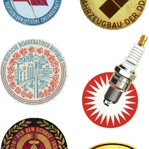 Op de afbeelding: Een verzameling van zes vintage ronde en ovale badges en stickers met verschillende ontwerpen. De badges tonen een rood-gele vlag met hamer en sikkel, een rood-witte cirkel met een stadsbeeld, een rood-witte cirkel met een bougie, een zwart-gouden cirkel met hamer en kompas, een bruine cirkel met een ruitvorm en de tekst "IFA", en een gele ovaal met de tekst "DDR".