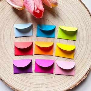 K&ouml;nnte beinhalten: Zehn kleine bunte Umschl&auml;ge mit abgerundeten Ecken, angeordnet in drei Reihen auf einer Holzoberfl&auml;che. Die Umschl&auml;ge sind in Blaut&ouml;nen, Gr&uuml;n, Gelb, Orange, Rot, Lila und Rosa.