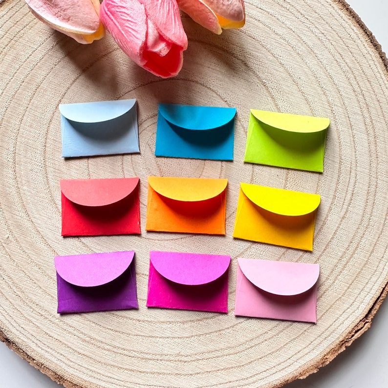 K&ouml;nnte beinhalten: Zehn kleine bunte Umschl&auml;ge mit abgerundeten Ecken, angeordnet in drei Reihen auf einer Holzoberfl&auml;che. Die Umschl&auml;ge sind in Blaut&ouml;nen, Gr&uuml;n, Gelb, Orange, Rot, Lila und Rosa.