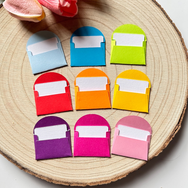 K&ouml;nnte beinhalten: Acht bunte Papierumschl&auml;ge mit wei&szlig;en Innenleben. Die Umschl&auml;ge sind in zwei Reihen zu je vier angeordnet, wobei sich die Farben abwechseln. Die Farben sind hellblau, rot, gelb, blau, rosa, lila, gr&uuml;n und orange.