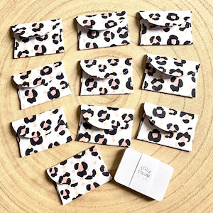 May include: Ten small white envelopes with a pink and black leopard print design. The envelopes are arranged in a circle on a wooden surface. A small white card with the text "Stella Stellina" is in the centre of the circle.