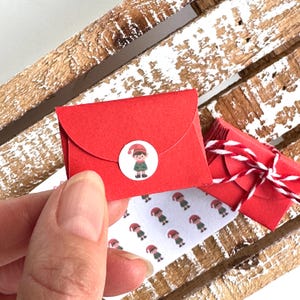 May include: A miniature red envelope with an elf sticker. Another small red package tied with red and white string is visible. A sheet of elf stickers is also present.