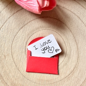 K&ouml;nnte beinhalten: Ein roter Umschlag mit einer wei&szlig;en Karte darin. Auf der Karte steht "Ich liebe dich" geschrieben, daneben ist ein Herz gezeichnet. Der Umschlag liegt auf einer Holzoberfl&auml;che.