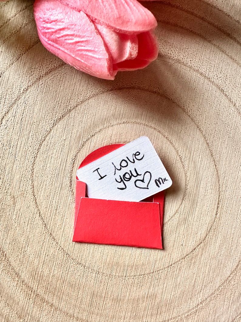 K&ouml;nnte beinhalten: Ein roter Umschlag mit einer wei&szlig;en Karte darin. Auf der Karte steht "Ich liebe dich" geschrieben, daneben ist ein Herz gezeichnet. Der Umschlag liegt auf einer Holzoberfl&auml;che.