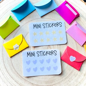 K&ouml;nnte beinhalten: Zwei S&auml;tze Mini-Aufkleber, jeder mit einem anderen Design. Der erste Satz hat einen wei&szlig;en Hintergrund mit dem Text "MINI STICKERS" und 12 Stern-Aufklebern. Der zweite Satz hat einen wei&szlig;en Hintergrund mit dem Text "MINI STICKERS" und 12 Herz-Aufklebern. Beide S&auml;tze sind von bunten Umschl&auml;gen umgeben.
