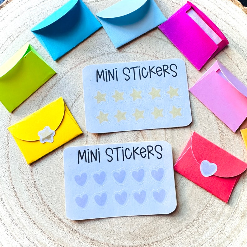 K&ouml;nnte beinhalten: Zwei S&auml;tze Mini-Aufkleber, jeder mit einem anderen Design. Der erste Satz hat einen wei&szlig;en Hintergrund mit dem Text "MINI STICKERS" und 12 Stern-Aufklebern. Der zweite Satz hat einen wei&szlig;en Hintergrund mit dem Text "MINI STICKERS" und 12 Herz-Aufklebern. Beide S&auml;tze sind von bunten Umschl&auml;gen umgeben.