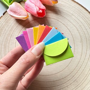 K&ouml;nnte beinhalten: Ein Satz bunter Papierumschl&auml;ge in einem Regenbogenmuster, gefaltet und f&auml;cherf&ouml;rmig in einer Hand ausgebreitet. Die Umschl&auml;ge sind klein und haben einen abgerundeten Verschluss.