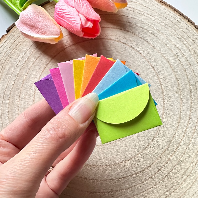 K&ouml;nnte beinhalten: Ein Satz bunter Papierumschl&auml;ge in einem Regenbogenmuster, gefaltet und f&auml;cherf&ouml;rmig in einer Hand ausgebreitet. Die Umschl&auml;ge sind klein und haben einen abgerundeten Verschluss.